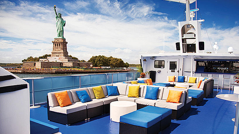 Open-air yacht lounge deck with colorful seating and Statue of Liberty views.